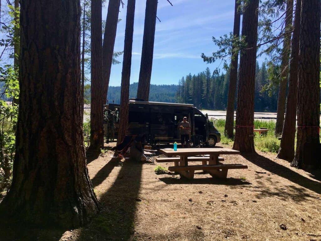 Camper van parked at campsite with tall trees and lake in the background