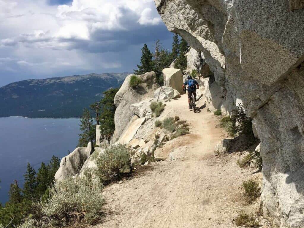 Mountain biker on narrow Tahoe Flume Trail with rock wall on one side and drop off on the other with Lake Tahoe below