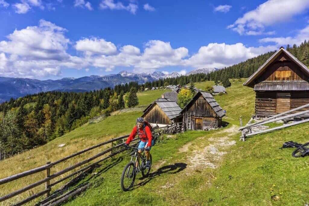 Mountain biker riding on trail in Slovenia with rustic mountain huts and mountains behind him