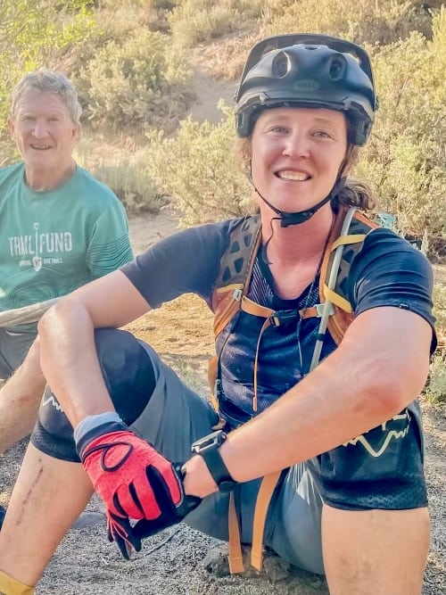 Becky, founder of the bike blog Two Wheeled Wanderer, sitting on ground wearing mountain biking gear with her dad behind her