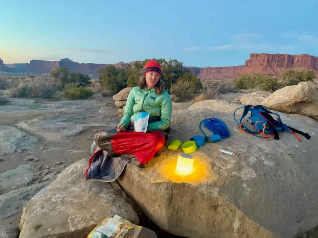 Woman bundled up in warm clothes while sitting on large rock eating dehyrated meal on the White Rim Trail in Utah