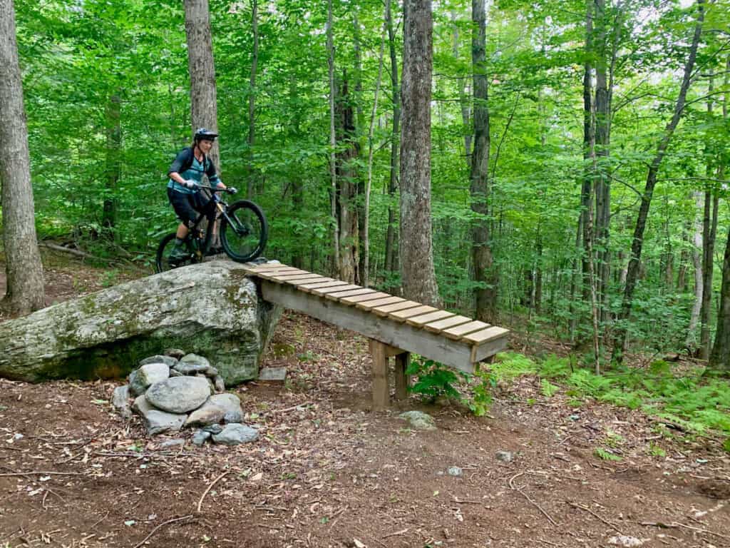 Mountain biker riding up and over rock roller onto elevated wooden ramp with drop off at end