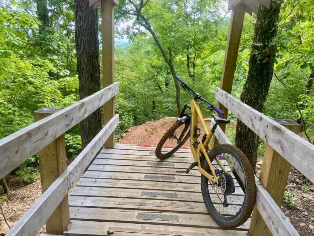 Mountain bike leaning against railing of wooden bridge leading to drop onto singletrack trail in Arkansas