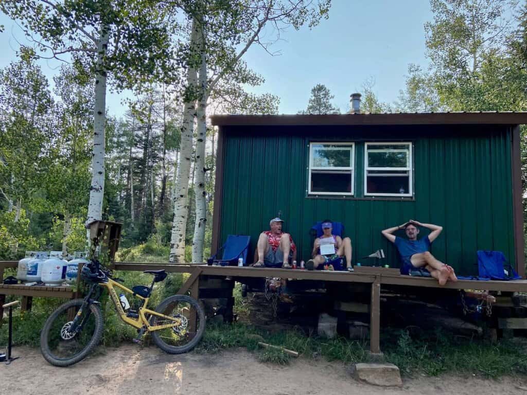 Three people sitting and relaxing on open porch in front of San Juan hut on the Telluride to Moab mountain bike route
