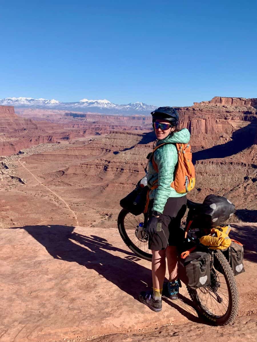 Becky smiling for photo next to loaded bikepacking bike on White Rim Trail in Utah with expansive views out over Canyonlands National Park
