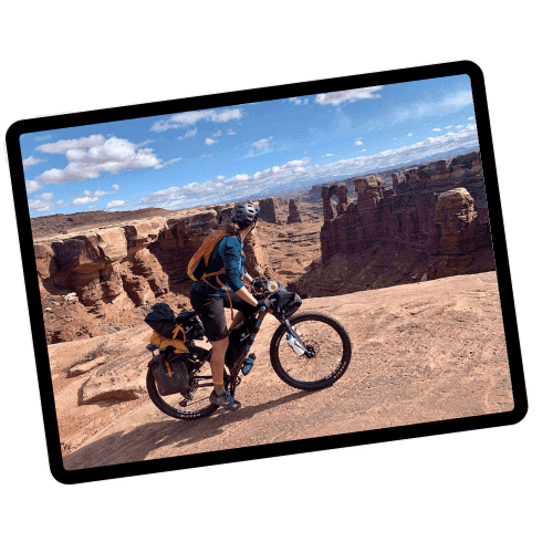 Bikepacker riding loaded bikpacking bike on rock ledge on White Rim Trail in Utah with Canyonlands views in distance. Photo is set in a tablet frame