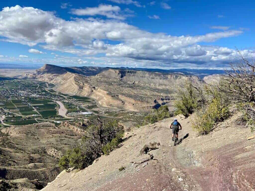 Mountain biker riding down narrow sidehill trail with drop off on right and epic Colorado mesa views in front