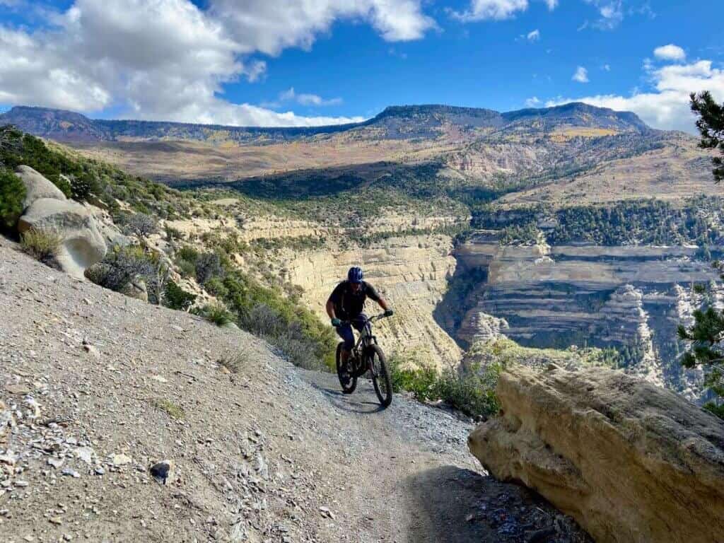 Mountain biker riding on singletrack trail with drop off on one side and high Colorado plateaus in background
