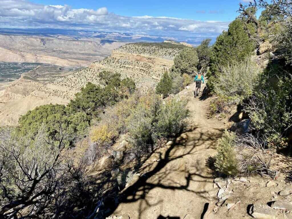 Mountain biker riding down narrow sidehill trail with drop off on right side and Colorado mesa views out front