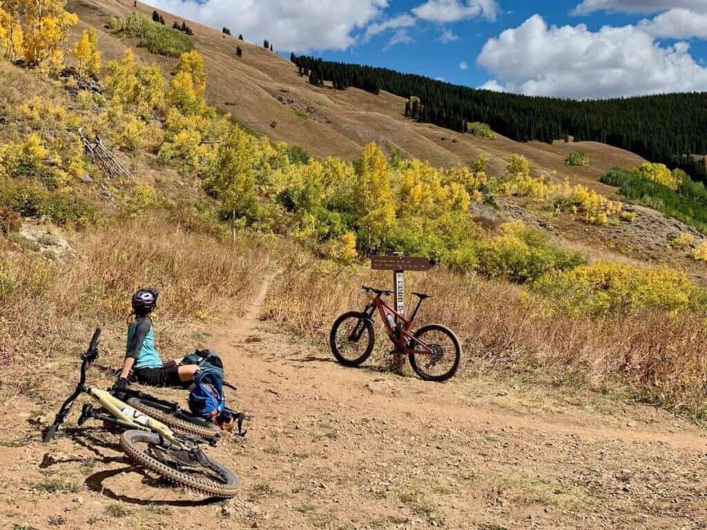 Mountain biker sitting on ground next to bikes looking out over changing aspen leaves landscapes on Teocalli Ridge Trail in Colorado