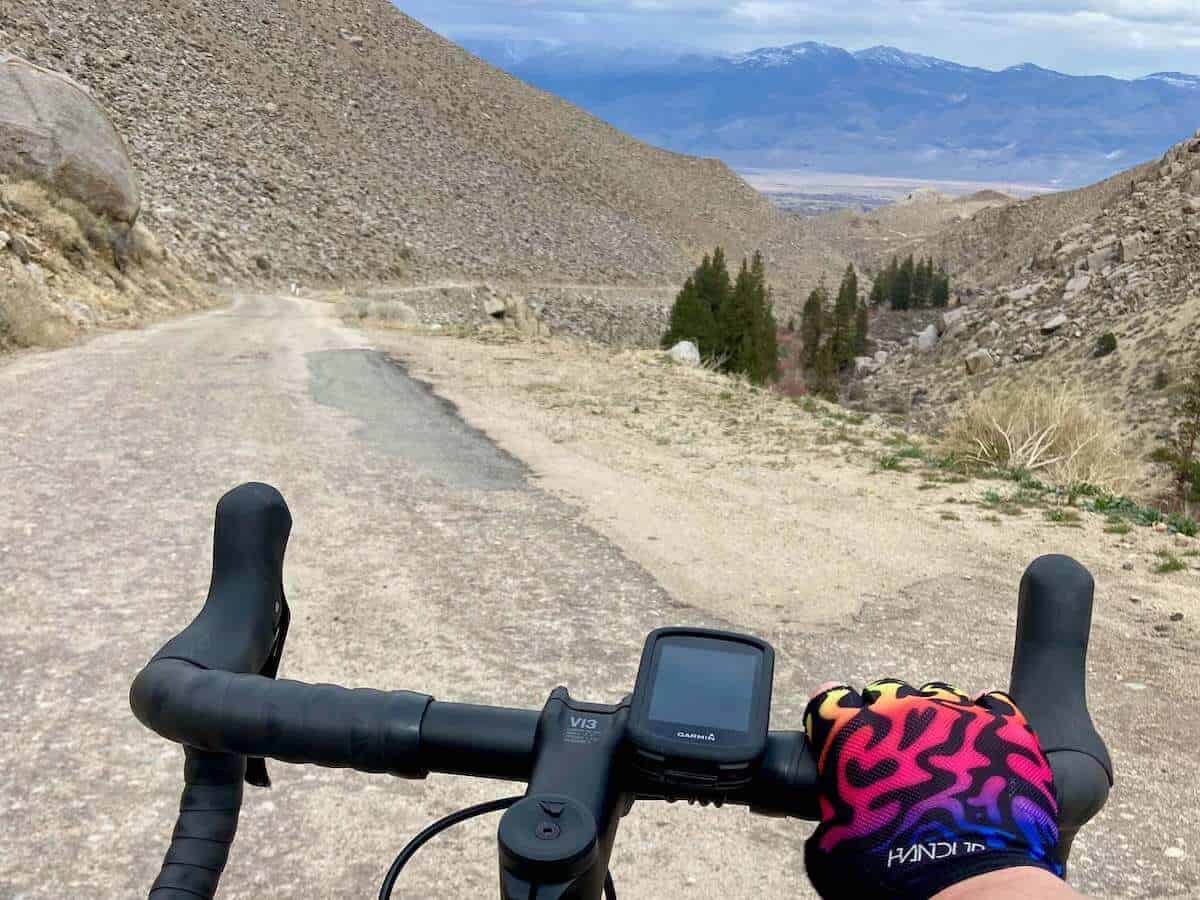 View out over road bike handlebar with Garmin Edge 830 mounted. Road winds through desert canyon with snow capped mountains in distance