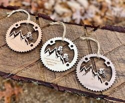 Three mountain bike Christmas ornaments lying next to each other shaped like a cog ring with mountain biker and mountains in the middle