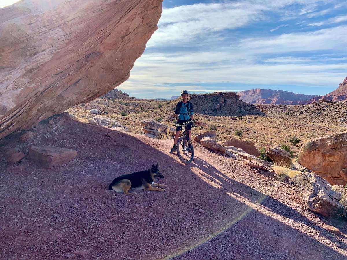 Mountain biker stopped on trail in Moab with dog laying down in shade under big boulder