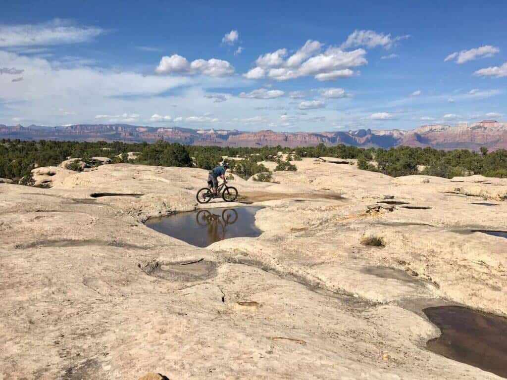 Mountain biker riding bike on large rock slab on Gooseberry Mesa in Utah with red bluffs in distance