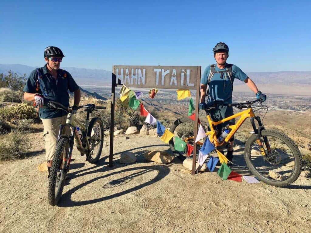 Two mountain bikers standing with bikes next to metal cut out sign at top of ridge that reads "Hahn Trail"