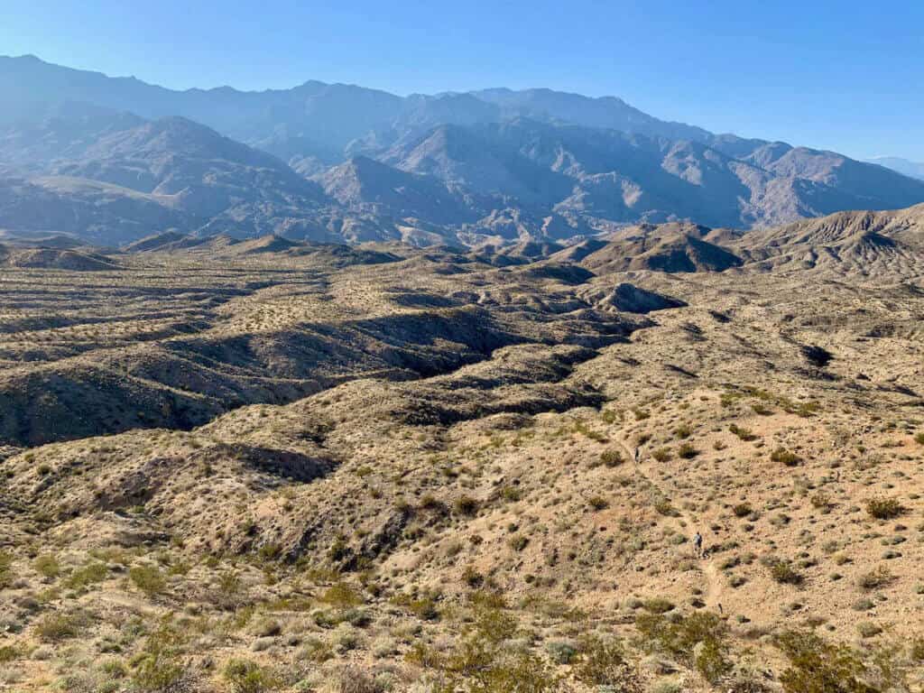 Landscape view of high desert mountains and ridges in California with faint singletrack trail below with two mountain bikers far away