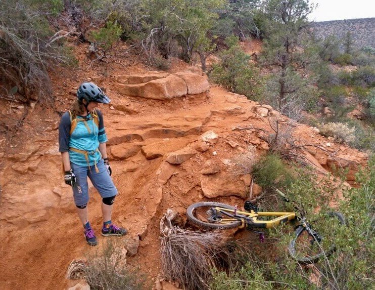 Woman standing on mountain bike trail in Sedona with mountain bike in the bushes below after minor crash