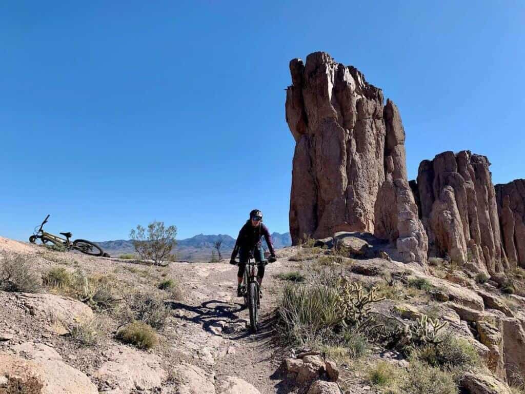 Mountain biker riding singletrack trail in desert of Kingman, Arizona with large rock monolith behind her