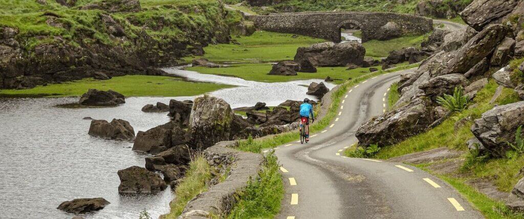 Cyclist on paved road next to water inlet in verdant green landscape of Ireland