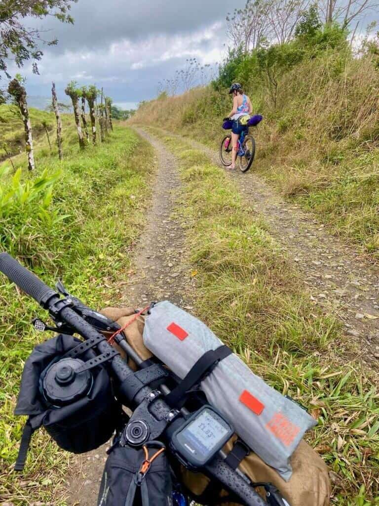 Photo out over front of bikepacking bike and handlebar roll of woman bikepacker stopped on remote dirt road