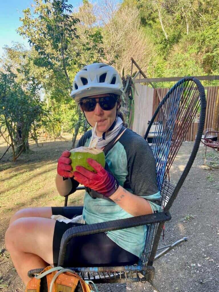 Becky sitting in chair drinking from a coconut with a straw wearing bike helmet and Ombraz sunglasses