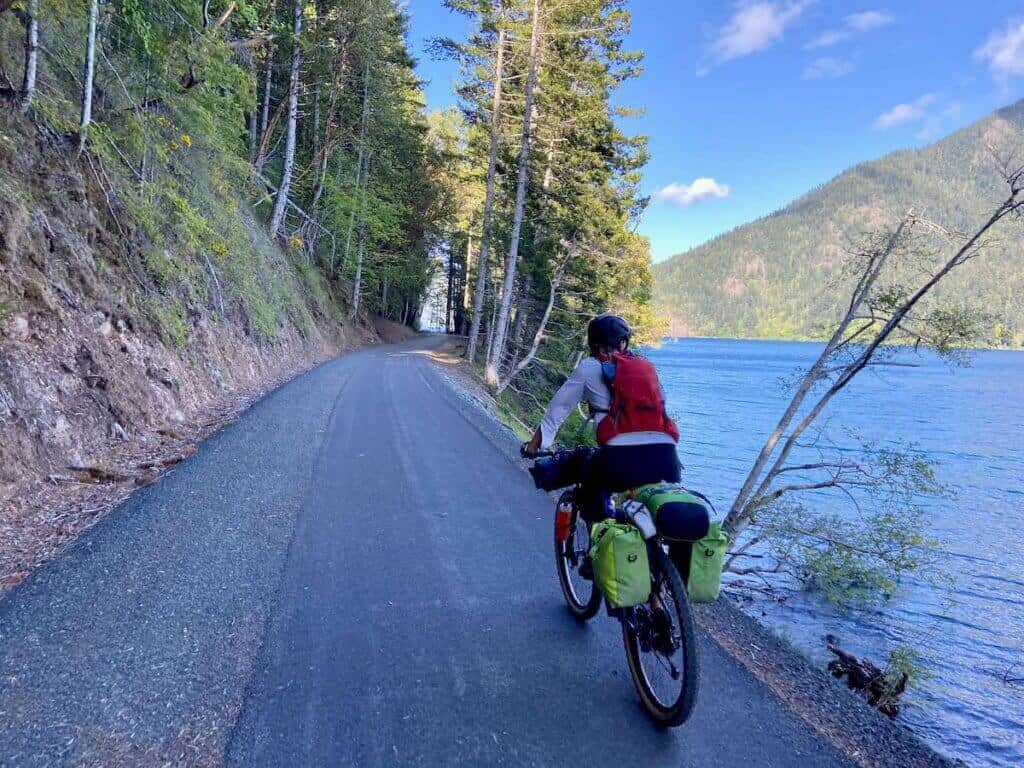 Woman riding loaded bikepacking bike on Olympic Discovery Trail in Washington with blue lake on righthand side