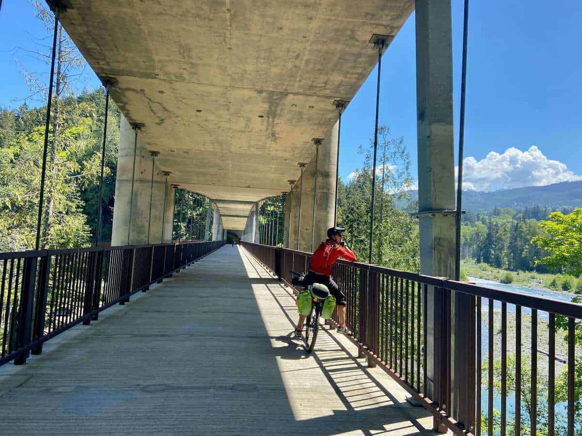 Woman straddling loaded bikepacking bike while looking over side of multi-use bridge walkway over river