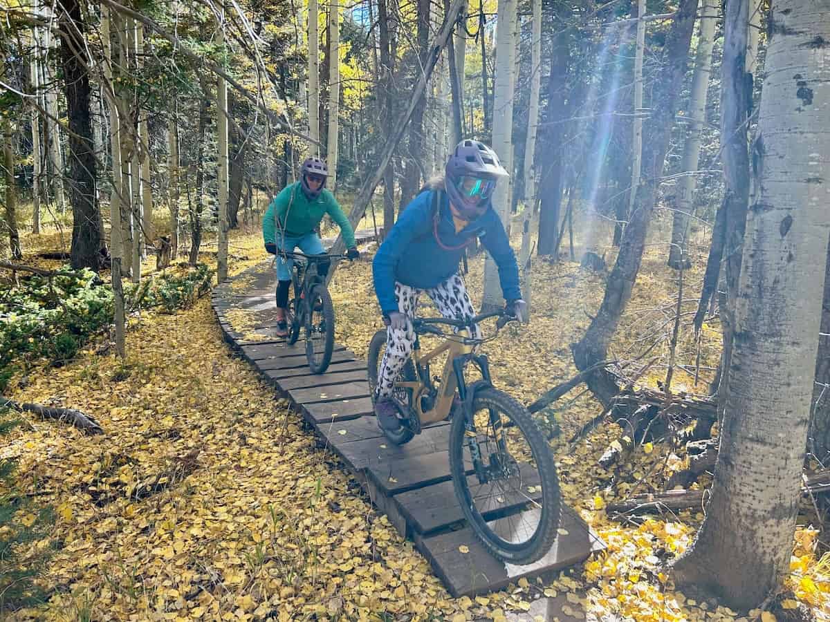 Two women riding mountain bikes over wooden bridge above downed golden fall foliage leaves at Angel Fire Bike Park in New Mexico