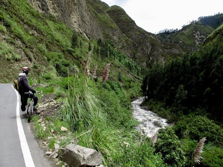 Cyclist stopped on bike by the side of road to take in the views of rushing river and green valley in Ecuador