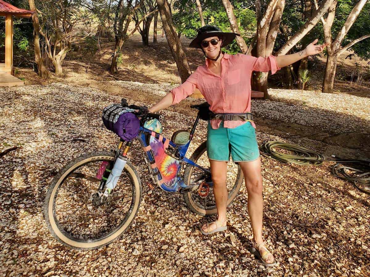 Woman standing next to loaded bikepacking bike with arms outstretched wide and smiling for camera