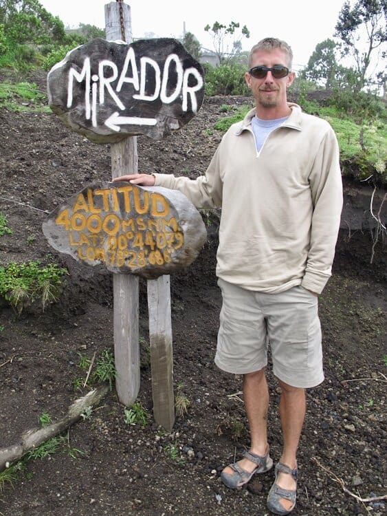 Man standing next to signs that read "Mirador" with arrow to the left and "altitud 2000 meters" on Cotopaxi Volcano in Ecuador