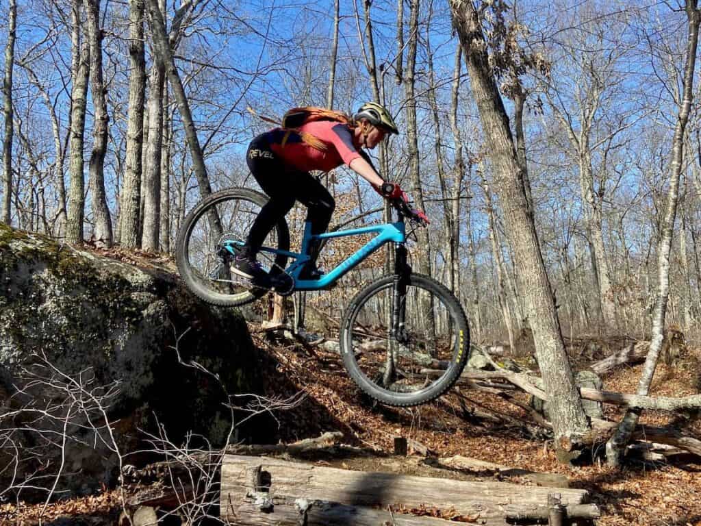 Becky riding mountain bike off of rock drop in woods wearing Revel Rider Flow pants
