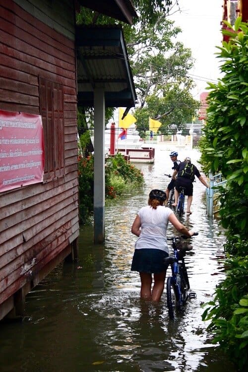 Three people walking bikes through flooded section of street in Thailand