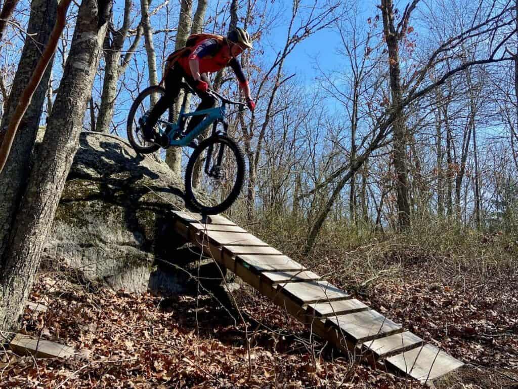 Female mountain biker riding bike down steep woody ramp off a rock at Woody Hill trail system in Rhode Island