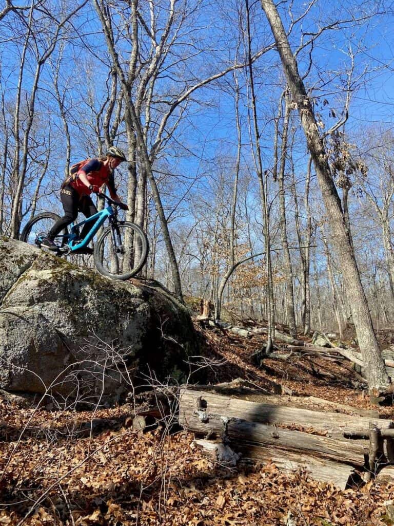 Becky akan mengendarai sepeda dari tebing batu setinggi 4 kaki di jaringan jalur Woody Hill di Rhode Island