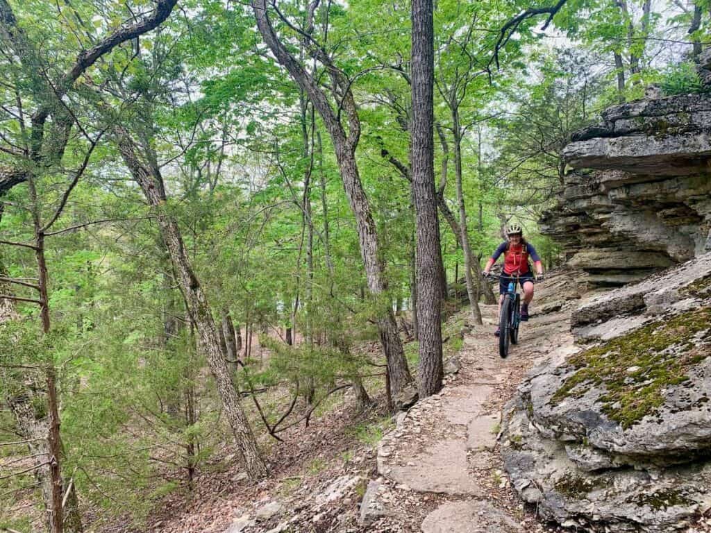 Mountain biker riding on narrow section of rocky trail with tall limestone cliff on left side of her at Hobbs State Park in Arkansas