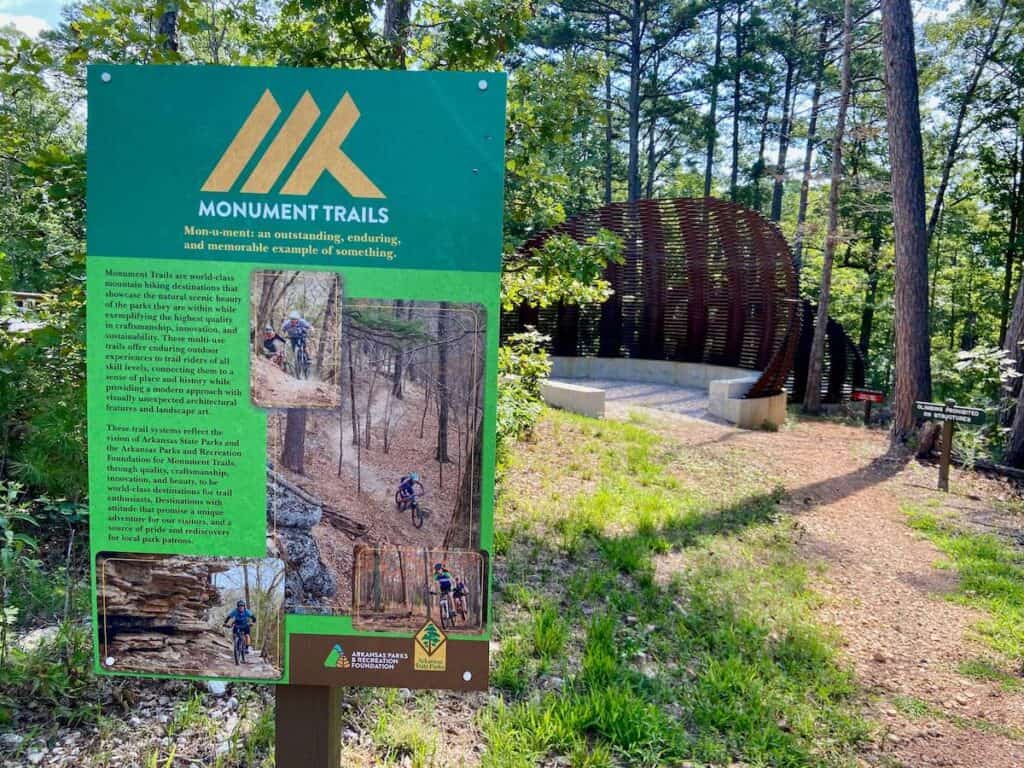Trail sign at Hobbs State Park in Arkansas describing what the Monument Trail System is. Large metal camping structures in background