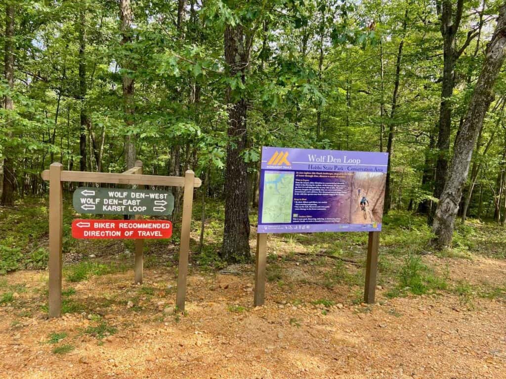 Trail signs at Hobbs State Park in Arkansas recommending route of travel for mountain bikers and trail names: Wolf Den-West (left), Wolf Den-East (right), Karst Loop (both directions)