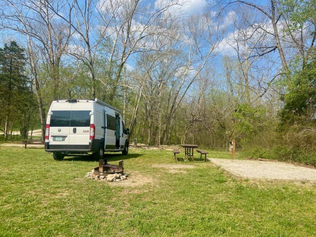 Van diparkir di padang rumput di sebelah meja piknik, cincin api, dan alas tenda berkerikil