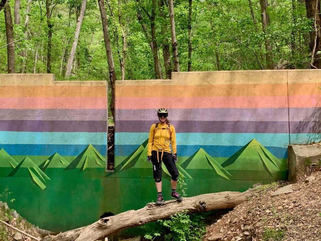Woman wearing mountain bike gear standing on fallen log in front of colorful mural on trail in Bentonville, Arkansas