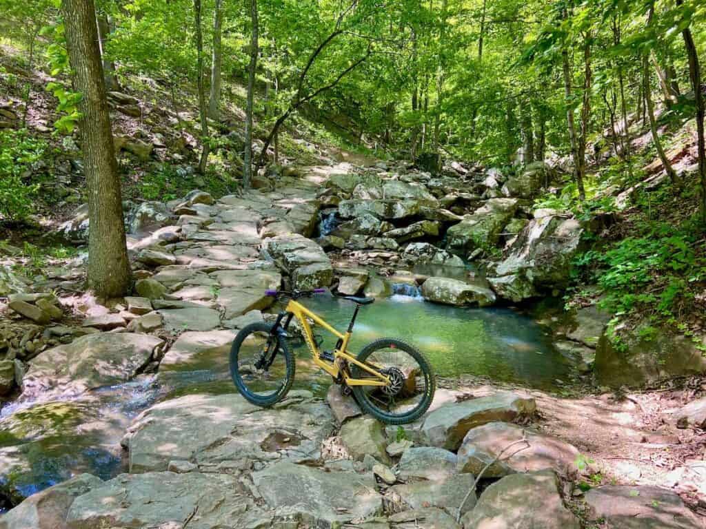 Mountain bike leaning against rock on rock-lined trail in a grotto with turquoise pool of water on trail in Mt. Nebo State Park in Arkansas