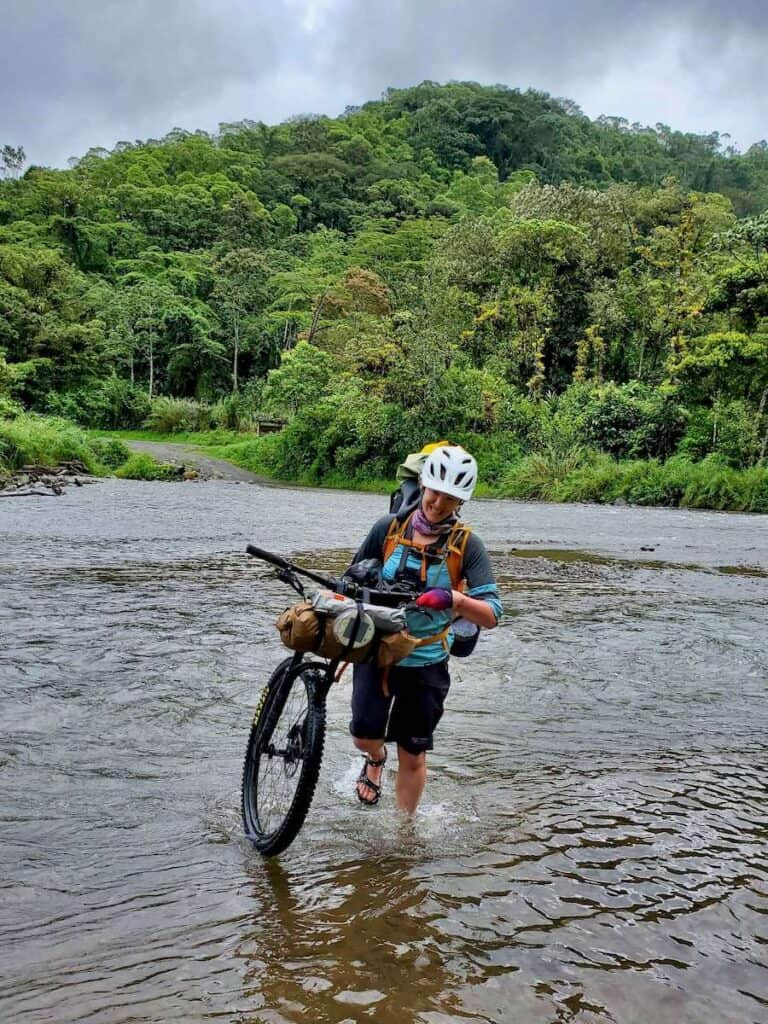 Woman carrying bike loaded with bikepacking bags across river in Costa Rica