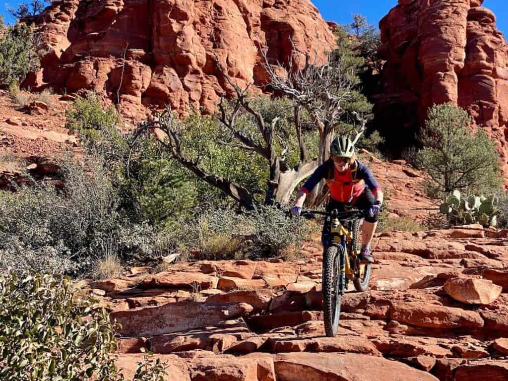 Becky riding mountain bike down steep, chunky section of red rock trail in Sedona