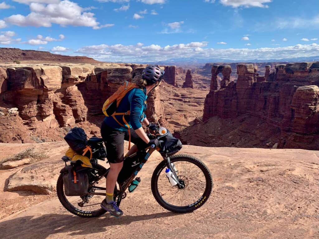 Bikepacking riding loaded bike next to edge of red slickrock edge with stunning Canyonland views next to her