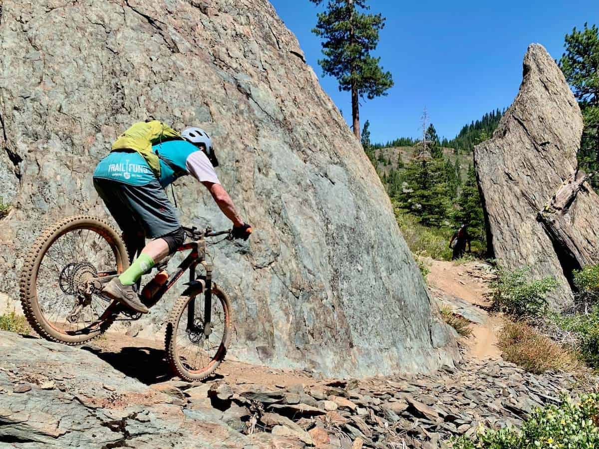 Man riding mountain bike down loose trail next to large rock boulder wearing bright green mountain bike socks