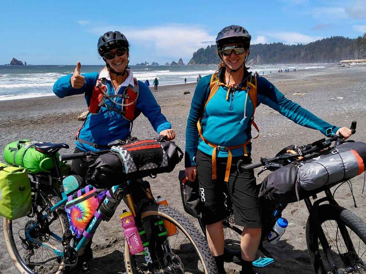 Two woman standing next to loaded bikepacking bikes on beach in Washington smiling for photo
