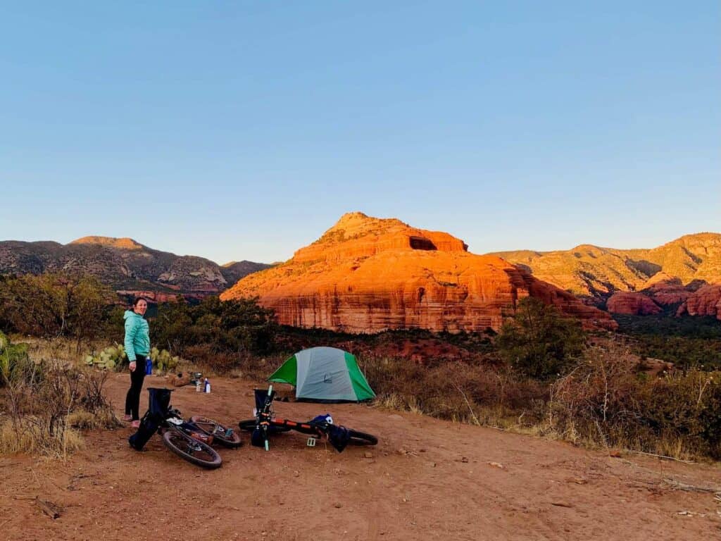 Woman standing next to dispersed campsite with tent set up and bikepacking bikes lying on ground. Setting sun is casting glow over surrounding bluffs outside of Sedona, Arizona