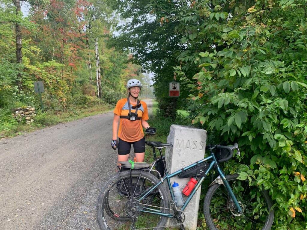Woman standing next to bikes leaning against stone state line boundary between Vermont and Massachusetts on the VTXL bikepacking route