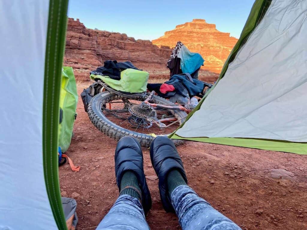 Photo from inside tent looking out onto woman's legs and feet wearing camp booties. Views of sun coming up over red rocks in Utah