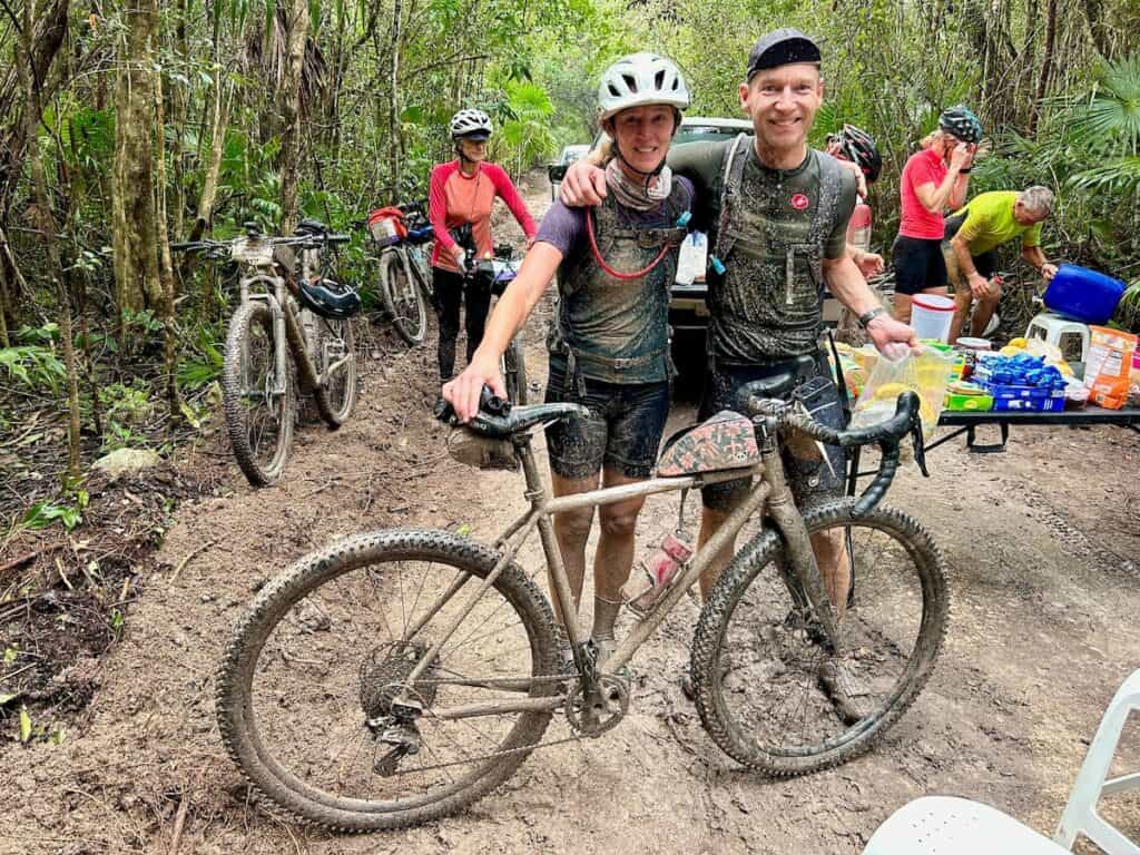 Two people posing for photo with bike and clothes caked in mud